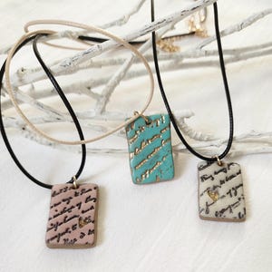 May include: Three rectangular pendant necklaces. One is light pink with black script and a gold heart. Another is turquoise with gold accents. The third is off-white with black script and a gold heart. Each pendant hangs from a cord.