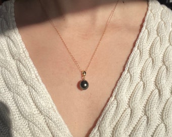 May include: A gold-colored chain necklace with a dark gray pearl pendant. The necklace is displayed against a white cable-knit sweater. The pearl is round and appears to be approximately 0.5 inches in diameter.