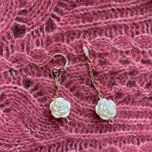 May include: A pair of gold-colored earrings with a floral design. Each earring features a small, white, rose-shaped pendant suspended from a delicate chain. The earrings have a hook closure and are adorned with small, pearl-like beads.