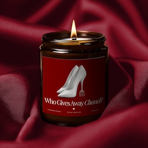 May include: A lit soy wax candle in a brown glass jar with a red label. The label features a graphic of two white high heels and the text "Who Gives Away Chanel?" The candle sits on a red satin surface.
