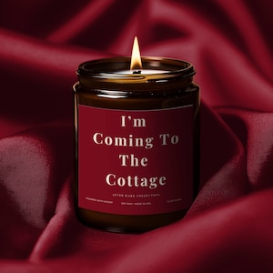 Puede incluir: Una vela encendida en un tarro de vidrio oscuro con una etiqueta roja. La etiqueta dice "I'm Coming To The Cottage" en texto blanco. La vela es parte de la "After Dark Collection" y está sobre un fondo de satén rojo oscuro.