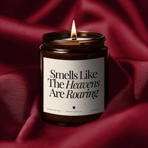 Puede incluir: Una vela de cera de soja encendida en un frasco de vidrio marrón con una etiqueta blanca. La etiqueta dice "Smells Like The Heavens Are Roaring". La vela está sobre una tela de satén rojo oscuro. Hecho en los EE. UU.
