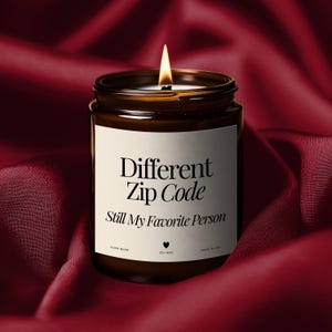 Könnte beinhalten: Eine brennende Sojawachskerze in einem braunen Glas mit einem weißen Etikett. Auf dem Etikett steht "Different Zip Code Still My Favorite Person". Die Kerze steht auf einem tiefroten Satinstoff.