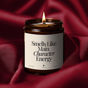 Op de afbeelding: Een brandende kaars in een donker amberkleurige glazen pot met een wit etiket met de zwarte tekst Smells Like Main Character Energy, en de tekst SLOW BURN, een hartje, SOY WAX en MADE IN USA onderaan het etiket gedrukt.