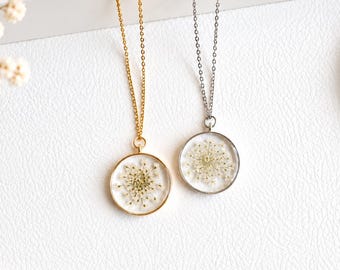 May include: Two round pendant necklaces featuring dried white flowers encased in clear resin. One pendant has a gold-toned metal frame and chain, while the other features a silver-toned metal frame and chain, both hanging vertically.