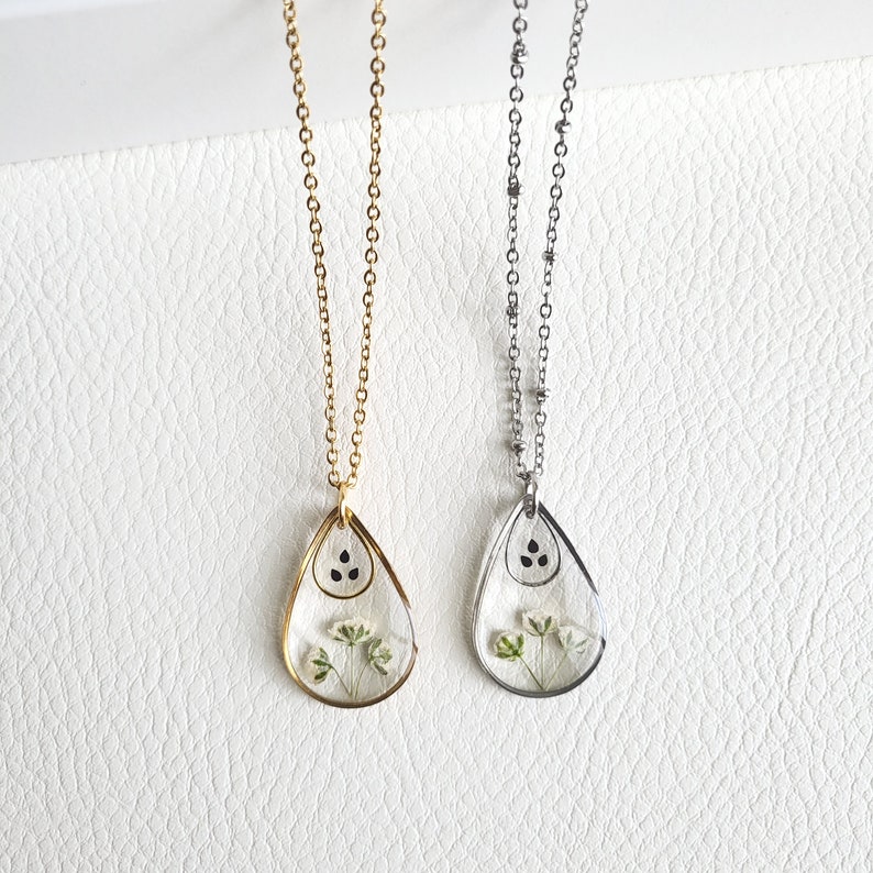 Pu&ograve; includere: Due delicate collane con ciondoli a forma di goccia che presentano fiori pressati e semi neri. Una collana ha una catena dorata e l'altra una catena argentata.
