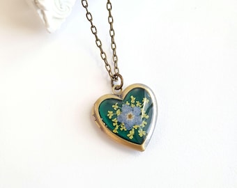 May include: A heart-shaped locket necklace with a bronze chain. The locket has a dark green background with a blue flower and yellow accents. The locket is made of metal and has a hinged opening.