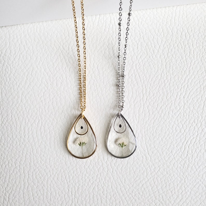 Pu&ograve; includere: Due delicate collane con ciondoli a forma di lacrima. Ogni ciondolo contiene un fiore bianco pressato e un piccolo punto nero. Una collana ha una catena dorata e l'altra una catena argentata.