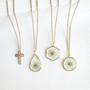 May include: Four gold-toned necklaces featuring clear resin pendants with embedded white pressed flowers. The pendants are shaped as a cross, a teardrop, a hexagon, and a circle, each suspended from a delicate gold-toned metal chain.