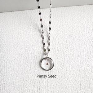 May include: A silver chain necklace with a clear oval pendant containing a single pansy seed. The pendant is hanging on a white background. The text "Pansy Seed" is written below the pendant.