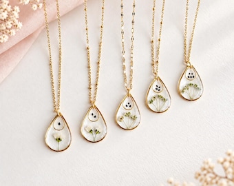 May include: Five gold necklaces featuring teardrop-shaped pendants. Each pendant contains clear resin with embedded dried white flowers and small black seeds. The necklaces have delicate gold chains and are arranged in a row.
