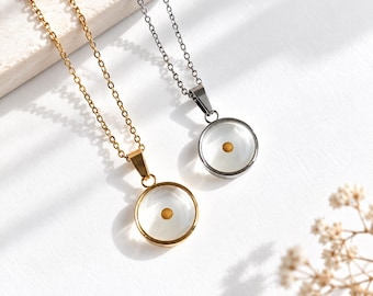 Puede incluir: Dos collares con colgante circular, uno en metal dorado y otro en metal plateado. Cada colgante presenta una carcasa circular transparente que contiene una única semilla pequeña de color mostaza suspendida en el centro del espacio.