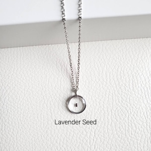 May include: A silver chain necklace with a small, round pendant. The pendant is clear and contains a tiny, dark seed inside. The text "Lavender Seed" is printed below the pendant.