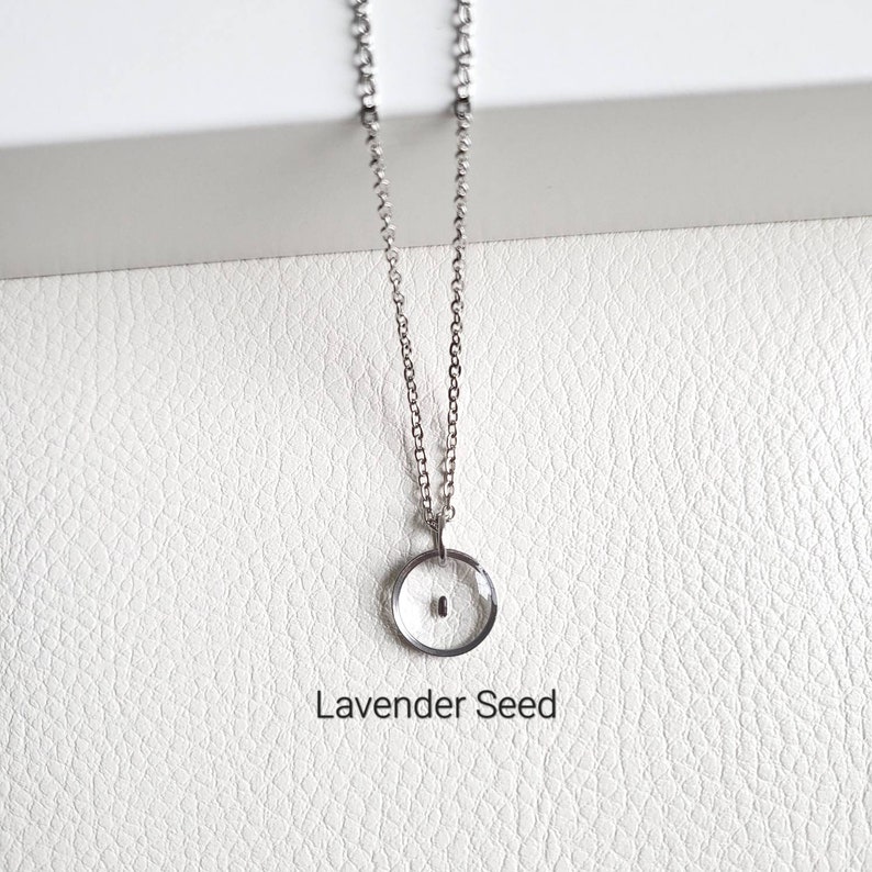 May include: A silver chain necklace with a small, round pendant. The pendant is clear and contains a tiny, dark seed inside. The text "Lavender Seed" is printed below the pendant.
