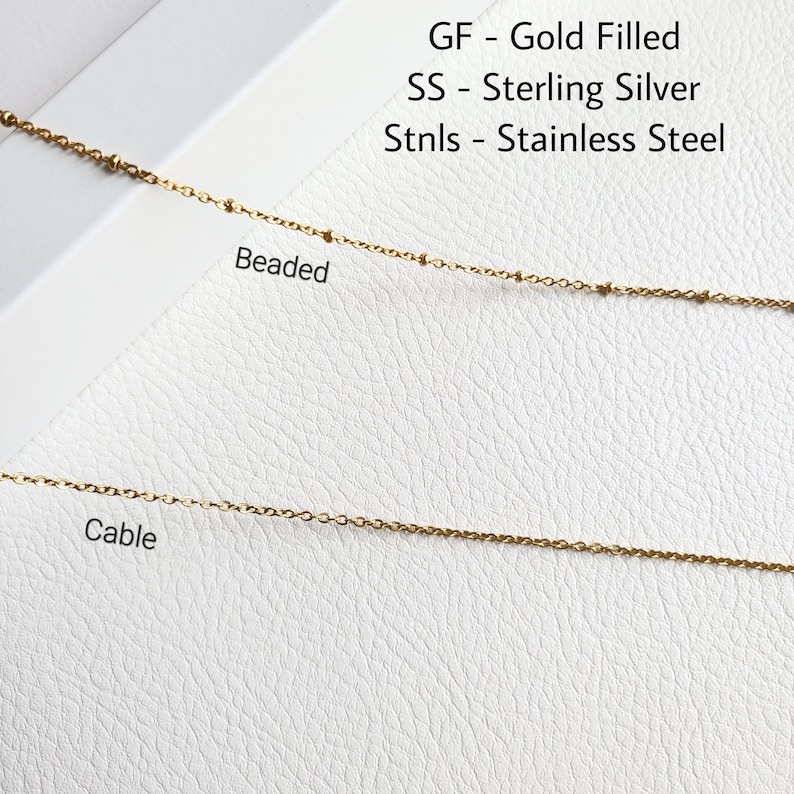 May include: Close-up of two gold-colored chain necklaces. The top necklace is beaded, and the bottom is a cable chain. Text in the image reads: GF - Gold Filled, SS - Sterling Silver, Stnls - Stainless Steel.