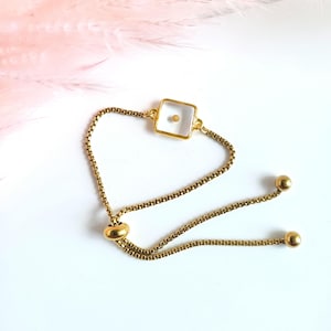 May include: Gold-toned adjustable bracelet featuring a square charm with a single seed. The bracelet has a delicate chain and two small ball accents. The background is white with a hint of pink.