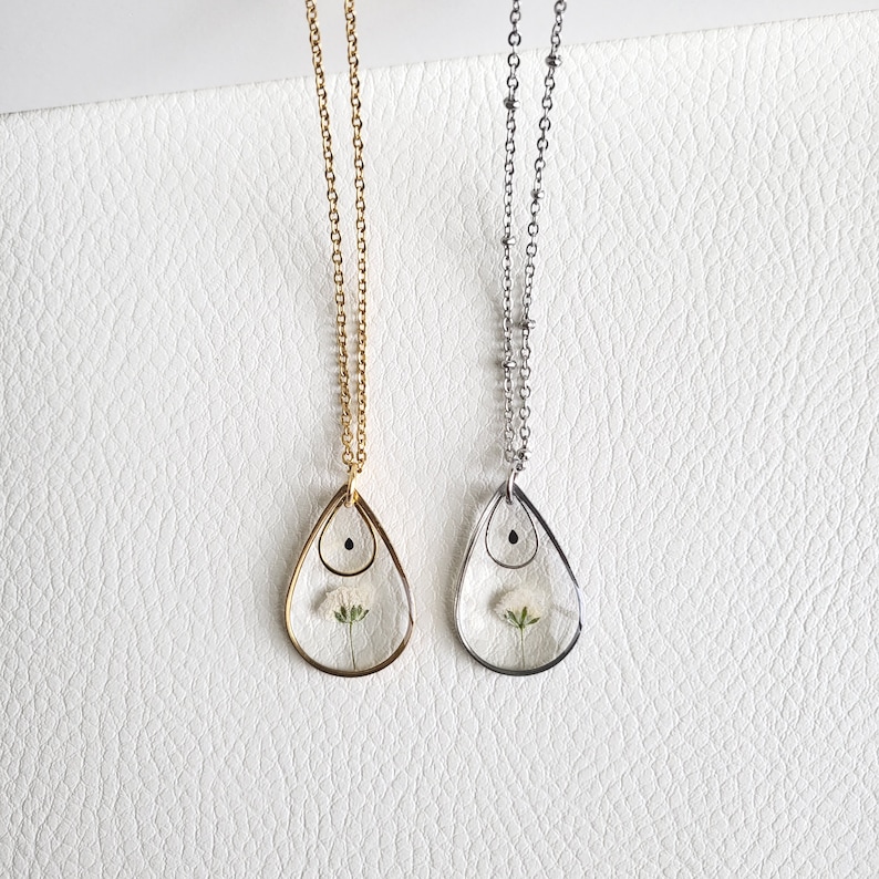May include: Two necklaces with teardrop pendants, one gold-toned and one silver-toned. Each pendant encases a small white flower. The chains are delicate, and the pendants have a small black accent at the top.