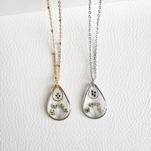 Pu&ograve; includere: Due delicate collane con ciondoli a forma di lacrima che presentano fiori pressati e semi neri. Le collane sono su uno sfondo bianco. Una collana ha una catena dorata e l'altra una catena argentata.