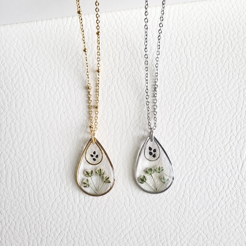 Pu&ograve; includere: Due delicate collane con ciondoli a forma di lacrima che presentano fiori pressati e semi neri. Le collane sono su uno sfondo bianco. Una collana ha una catena dorata e l'altra una catena argentata.