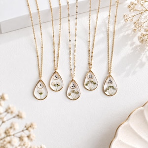 May include: Five gold necklaces featuring teardrop-shaped pendants with clear resin centers. Each pendant contains pressed white flowers and small dark seeds or petals. The necklaces have delicate gold chains of varying link styles.