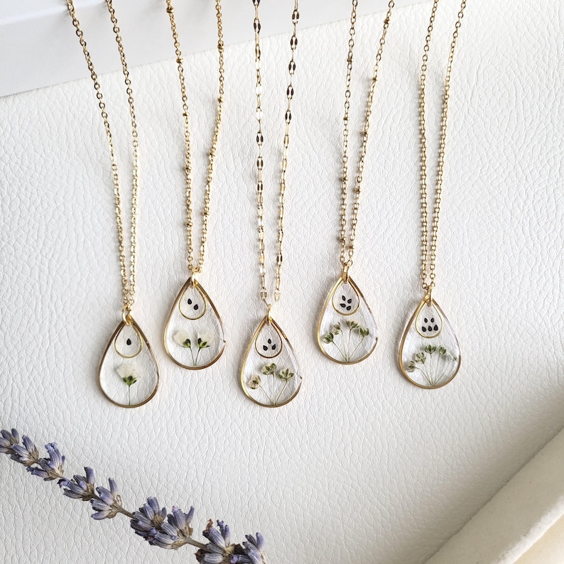 Pu&ograve; includere: Cinque collane d'oro con catene delicate e ciondoli a forma di goccia. Ogni ciondolo presenta un design in resina trasparente con fiori secchi pressati e semi neri.