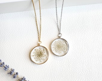 May include: Two delicate necklaces with round pendants featuring pressed white flowers. One necklace has a gold chain and the other has a silver chain.