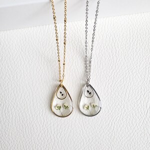Pu&ograve; includere: Due delicate collane con ciondoli a forma di lacrima che presentano fiori bianchi pressati e semi neri. Le collane sono su uno sfondo bianco. Una collana ha una catena dorata e l'altra una catena argentata.