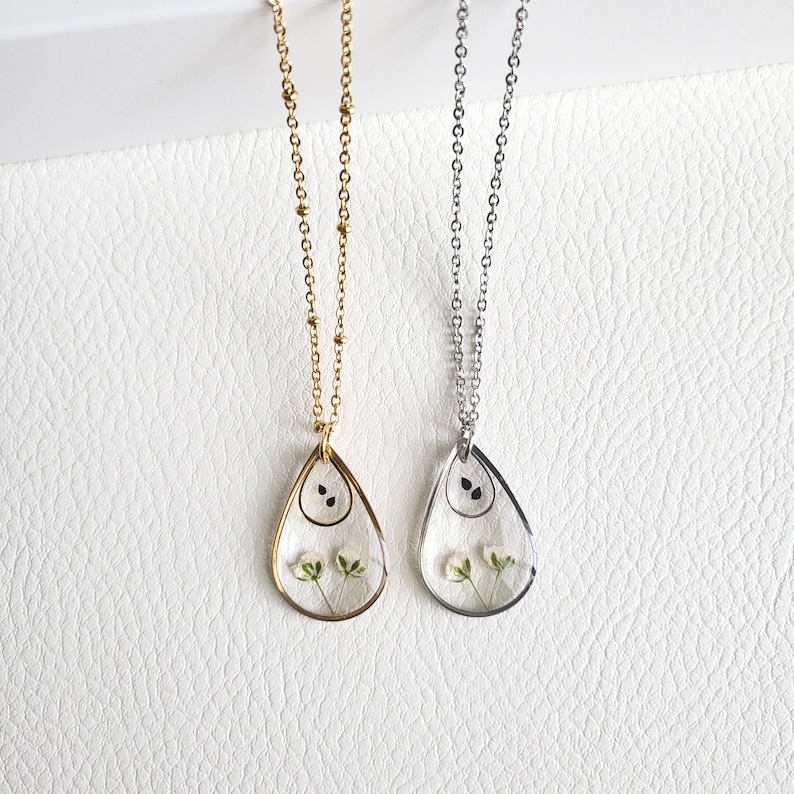 Pu&ograve; includere: Due delicate collane con ciondoli a forma di lacrima che presentano fiori bianchi pressati e semi neri. Le collane sono su uno sfondo bianco. Una collana ha una catena dorata e l'altra una catena argentata.