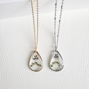 May include: Two necklaces with teardrop pendants. One necklace has a gold chain and pendant frame, the other has a silver chain and pendant frame. Each pendant contains dried flowers and black dots.