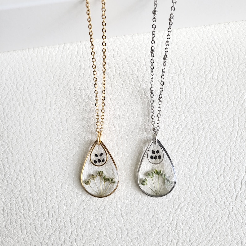 May include: Two necklaces with teardrop pendants. One necklace has a gold chain and pendant frame, the other has a silver chain and pendant frame. Each pendant contains dried flowers and black dots.