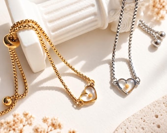 Può includere: Due braccialetti regolabili con catena a maglia quadrata, uno color oro e uno color argento. Ogni braccialetto presenta un ciondolo centrale a forma di cuore aperto contenente una piccola perlina gialla. Includono un meccanismo scorrevole.