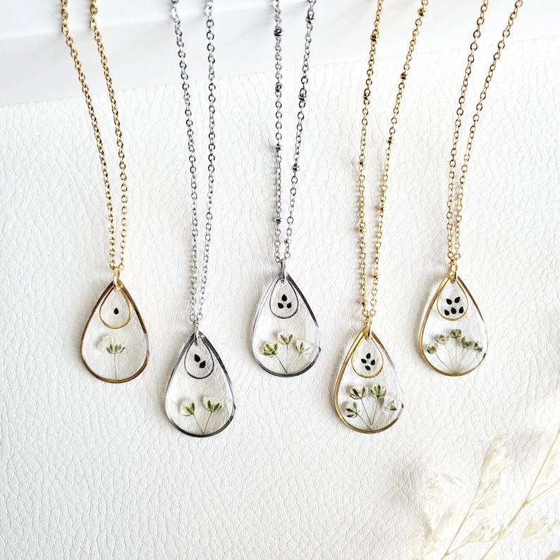 May include: Five teardrop-shaped pendant necklaces. Each pendant features a clear resin design with dried flowers and a small black accent. The necklaces have gold and silver chains.