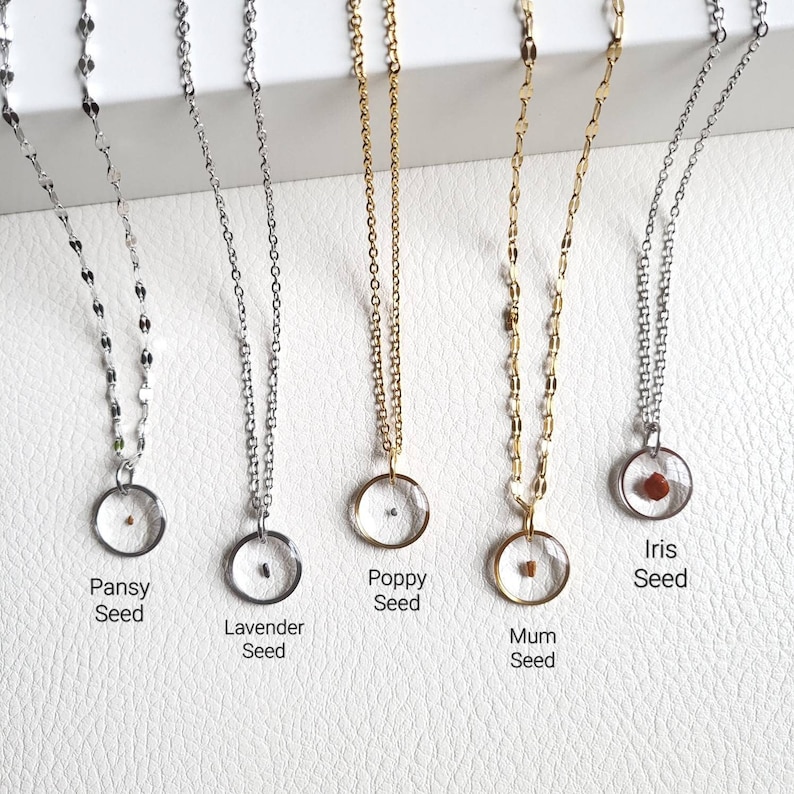 May include: Five silver and gold necklaces with clear glass pendants containing different flower seeds. The pendants are labeled "Pansey Seed", "Lavender Seed", "Poppy Seed", "Mum Seed", and "Iris Seed".