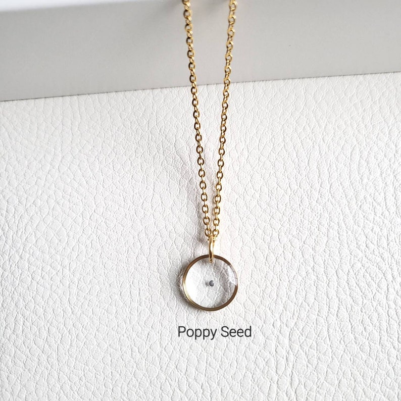 May include: A gold chain necklace with a clear glass pendant containing a poppy seed. The pendant is round and has a gold rim. The text "Poppy Seed" is written below the pendant.