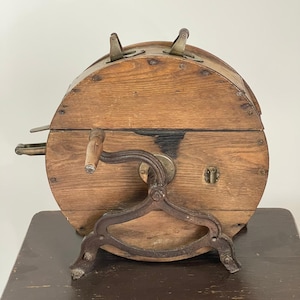 May include: Antique wooden butter churn with a metal crank and stand. The churn is barrel-shaped with a hinged lid and metal latches. The wooden body shows signs of age and wear, with a dark stain on the front.