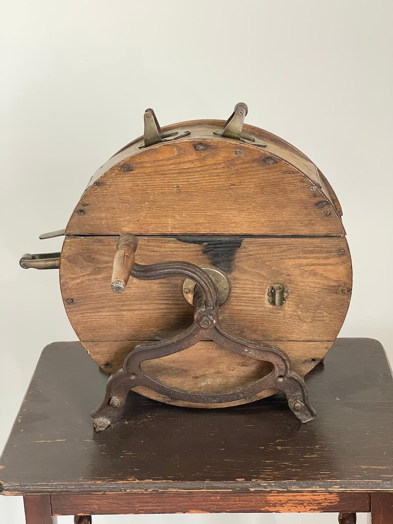 May include: Antique wooden butter churn with a metal crank and stand. The churn is barrel-shaped with a hinged lid and metal latches. The wooden body shows signs of age and wear, with a dark stain on the front.
