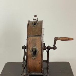 May include: An antique wooden and metal lottery wheel with a hand crank. The wheel is made of wood and has metal accents. The base is metal and has the word "IMPROVED" on it. The wheel is sitting on a dark wooden table.