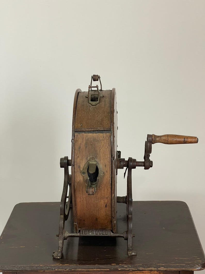 May include: An antique wooden and metal lottery wheel with a hand crank. The wheel is made of wood and has metal accents. The base is metal and has the word "IMPROVED" on it. The wheel is sitting on a dark wooden table.