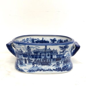 Könnte beinhalten: Ein blau-weißer Keramik-Pflanztopf mit rechteckiger Form und zwei Griffen. Der Pflanztopf zeigt eine detaillierte Landschaftsszene mit Gebäuden, Bäumen und Figuren. Der Rand und die Griffe sind in einem dunkleren Blauton gehalten.