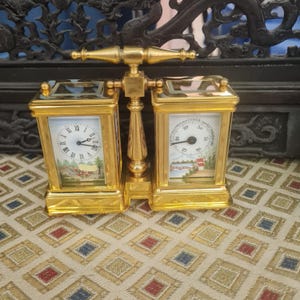 Könnte beinhalten: Eine goldfarbene, doppelseitige Uhr mit einem römischen Zifferblatt auf der einen Seite und einem Thermometer auf der anderen. Das Zifferblatt hat eine gemalte Szene mit einer Pferdekutsche. Das Thermometer hat eine gemalte Szene mit einer Brücke.