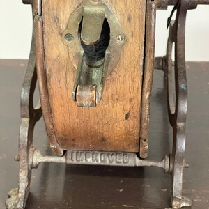 May include: Close-up of an antique wooden and metal object. The wooden portion is rectangular with a brass fitting and a dark opening. The metal frame is dark and has the word "IMPROVED" stamped on it. The object appears to be a vintage tool.