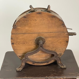 May include: Antique wooden lottery wheel with metal accents. The wheel is cylindrical, made of light brown wood, and features metal hinges and a crank. It rests on a decorative metal stand with claw feet, showcasing a vintage aesthetic.