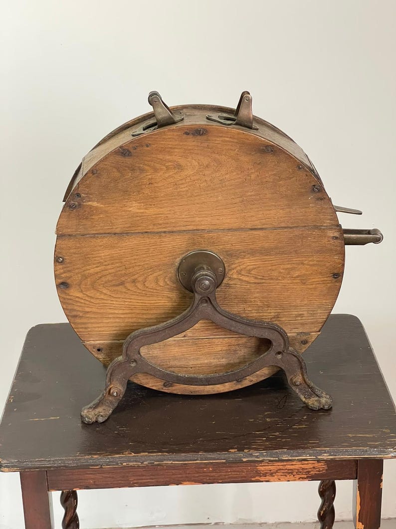 May include: Antique wooden lottery wheel with metal accents. The wheel is cylindrical, made of light brown wood, and features metal hinges and a crank. It rests on a decorative metal stand with claw feet, showcasing a vintage aesthetic.