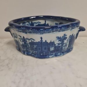 Può includere: Una fioriera in ceramica blu e bianca con un disegno paesaggistico. La fioriera ha un bordo smerlato e due piccole maniglie. Il design presenta edifici, alberi e figure in uno stile classico e vintage.