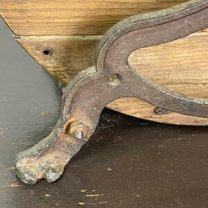 May include: Close-up of a vintage wooden furniture piece with a metal leg. The leg is ornate, with a claw foot design and a rusty, aged appearance. The wood has a natural grain and a warm, light brown color.