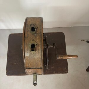 May include: An antique wooden pencil sharpener with a hand crank and a wooden handle. The sharpener is made of wood and metal, and it sits on a dark brown wooden table. The sharpener has two metal slots and a metal lever.