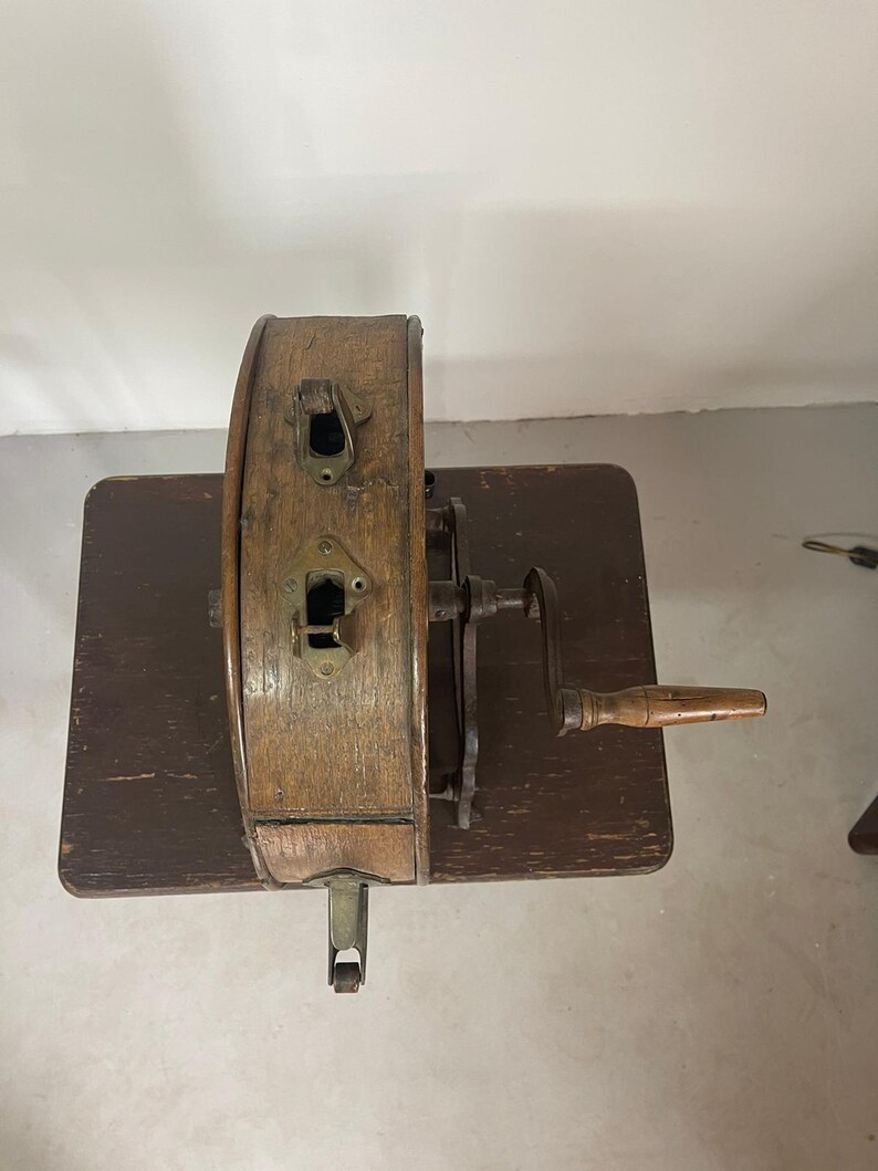 May include: An antique wooden pencil sharpener with a hand crank and a wooden handle. The sharpener is made of wood and metal, and it sits on a dark brown wooden table. The sharpener has two metal slots and a metal lever.