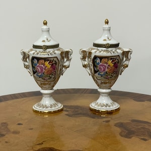 Może przedstawiać: Para białych porcelanowych urn z pokrywkami, ozdobionych złotymi wykończeniami, ozdobnymi uchwytami oraz centralnymi panelami z kolorowymi bukietami kwiatów. Każda urna posiada podstawę na cokole i szpiczasty uchwyt na pokrywce z zawiasami.