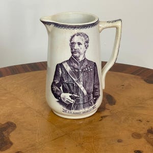 Może przedstawiać: Kremowy ceramiczny dzban z fioletowym portretem mężczyzny w mundurze wojskowym. Dzbanek ma uchwyt i ozdobną fioletową obwódkę. Tekst "SIR GARNET WOLSELEY" znajduje się na dole.