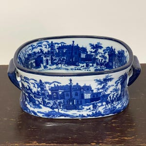 Könnte beinhalten: Eine ovale Keramikschale mit einem blau-weißen Transferdruck-Muster, das eine Landschaft mit Gebäuden, Figuren und Pferden zeigt. Die Schale hat zwei dunkelblaue Seitengriffe und ein durchgehendes Motiv auf der Außen- und Innenseite.